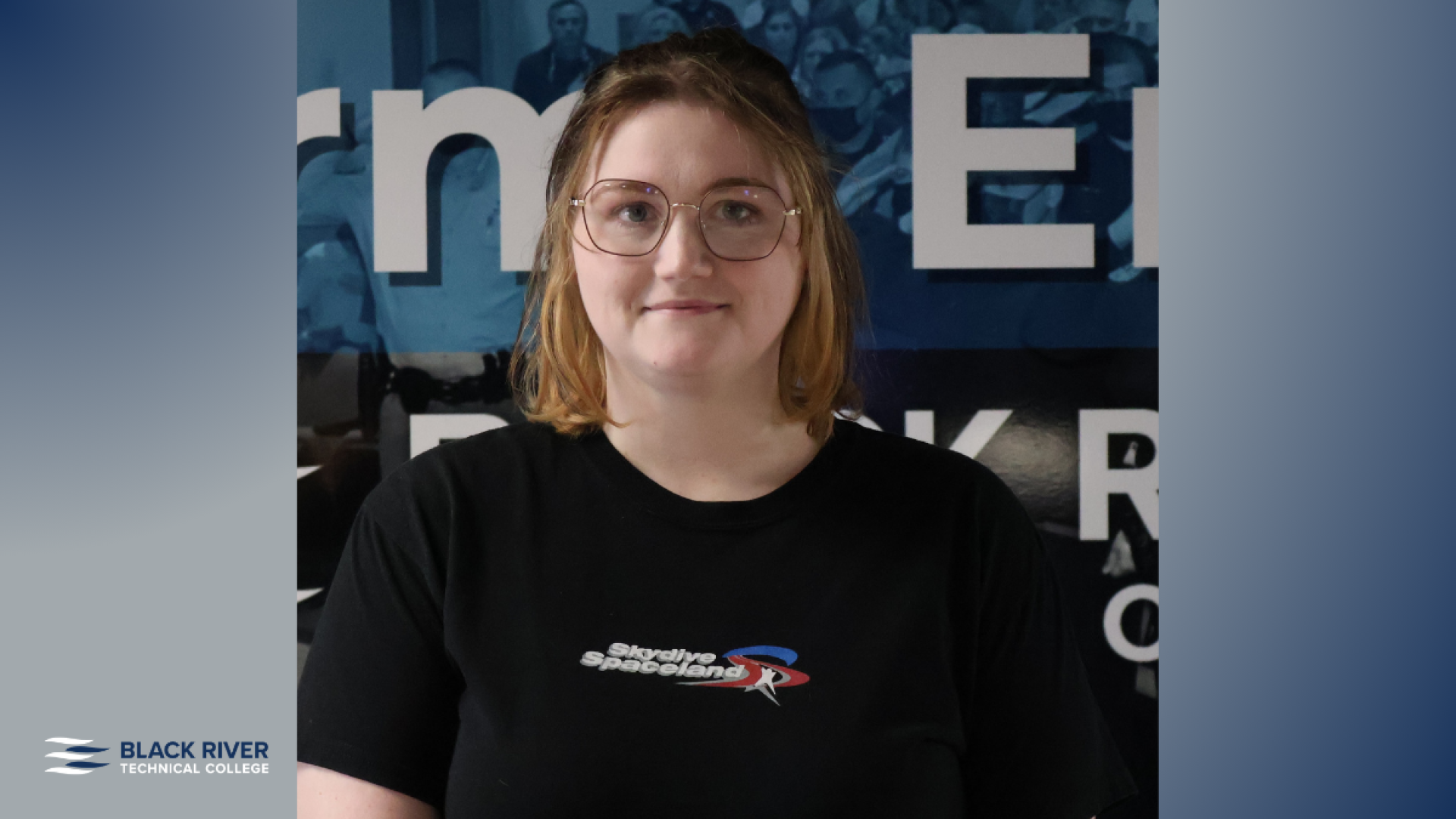 A young woman with shoulder-length reddish-blonde hair and glasses, wearing a black t-shirt, smiles in front of a Black River Technical College banner.
